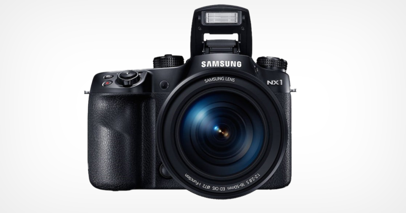 A Samsung NX1 camera is shown from the front with its lens in focus. The built-in flash is popped up, and the camera displays a textured grip on its body. The lens reflects a slight blue tint.