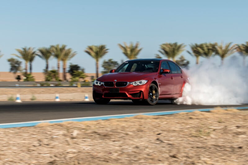 A red sports car drifts on a track, kicking up a cloud of smoke. Palm trees and a clear blue sky are in the background.