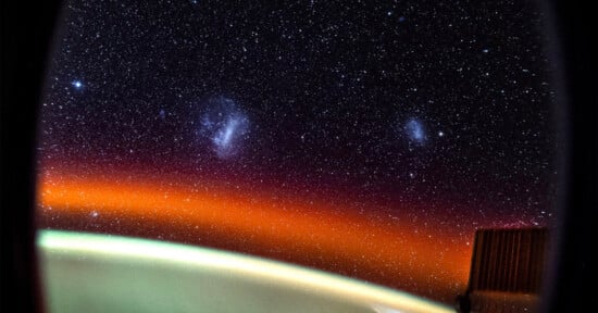 View of a starry sky from space, showcasing two prominent fuzzy galaxies against a dark backdrop. A warm orange glow and part of a spacecraft frame the lower portion of the scene.