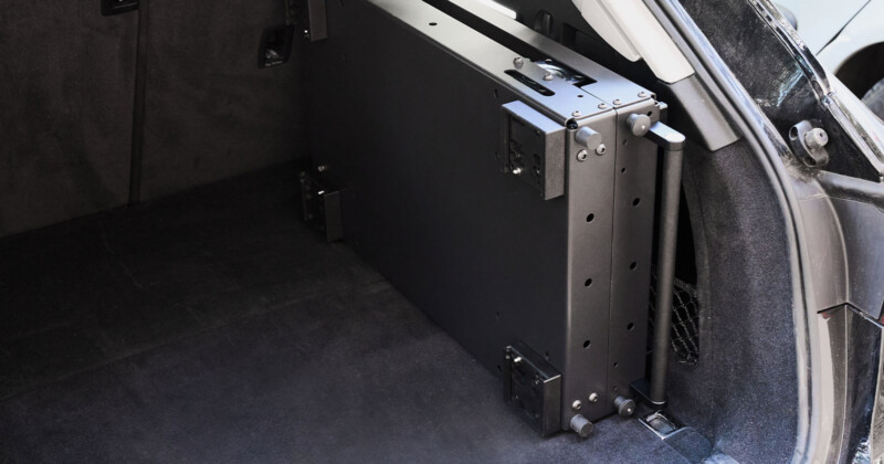 A close-up of a car's trunk showing a large, rectangular, black folding table neatly secured inside. The trunk space is carpeted, with visible side panels and netting for additional storage.