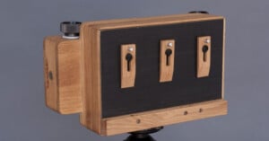 Wooden pinhole camera mounted on a tripod. The camera has a rectangular shape with three pinholes on the front, using a combination of light and dark wood finishes.