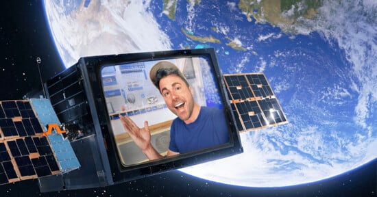 A man smiling and gesturing inside a screen on a satellite, with Earth visible in the background.