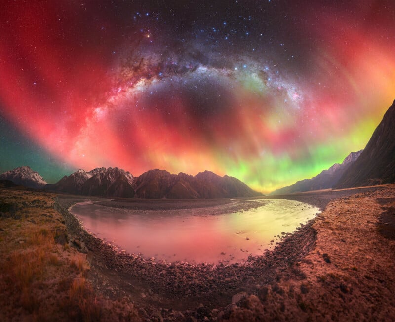 A vibrant night sky displaying colorful auroras and the Milky Way arches over a serene, reflective lake. Snow-capped mountains and rocky terrain create a stunning natural landscape under the celestial display.