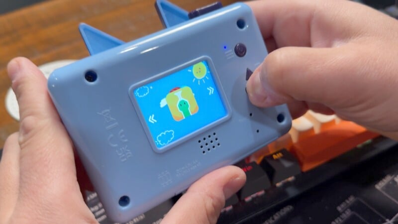 A person holds a small, blue handheld gaming device with animal ears. The screen displays a colorful game featuring a character with arrows indicating movement options.