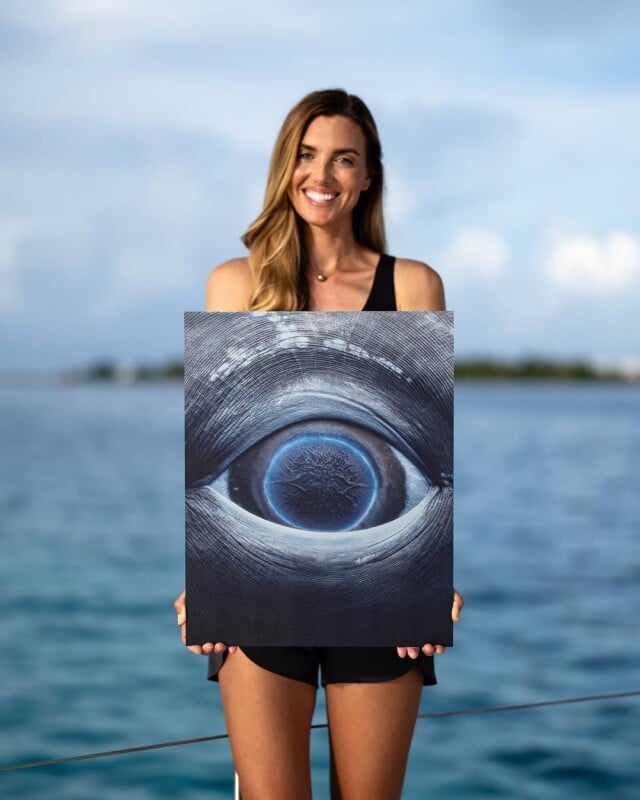 A woman stands on a boat holding a piece of art depicting a blue eye. The ocean is in the background under a partly cloudy sky. She is smiling and wearing a black sleeveless top and shorts.