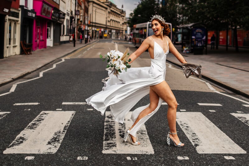 A joyful bride wearing a flowing white dress and silver heels runs across a city street. She's holding a bouquet and a purse, with a backdrop of colorful buildings and trees. The scene captures a sense of excitement and movement.