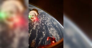 A spacecraft approaches a space station over Earth, with a view of clouds and the planet's surface below. The craft is illuminated by red and green lights, and parts of the station are visible in the foreground.