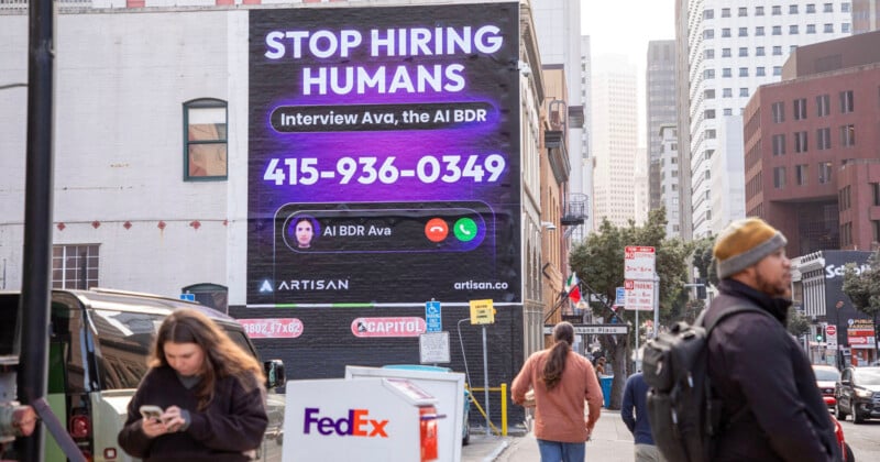 A busy city street features a large advertisement on a building. The ad reads "STOP HIRING HUMANS. Interview Ava, the AI BDR" with a phone number and an image of a virtual assistant interface. Pedestrians and cars are in the foreground.