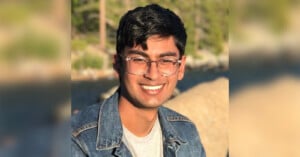 A person wearing glasses and a denim jacket smiles while sitting outdoors. Trees and water are blurred in the background, suggesting a natural setting.