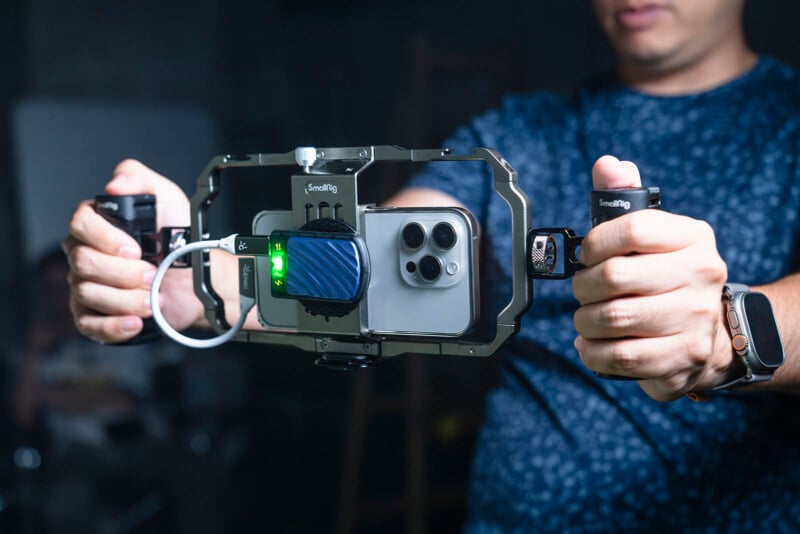 A person holding a smartphone stabilizer rig with a smartphone mounted on it. The rig has multiple attachments, including a microphone and cables. The person is wearing a smartwatch and a blue patterned shirt.