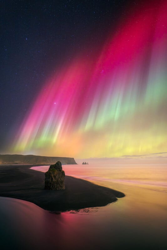 Colorful aurora borealis illuminates the night sky over a black sand beach. A rock formation stands in the foreground, with vibrant red, green, and yellow lights reflecting on the water. A clear starlit sky completes the scene.