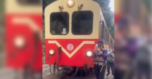 A vintage train with a red and beige color scheme travels along tracks surrounded by lush greenery. A person wearing a blue hat and holding walking poles walks near the train. The scene is outdoors, suggesting a day of hiking.