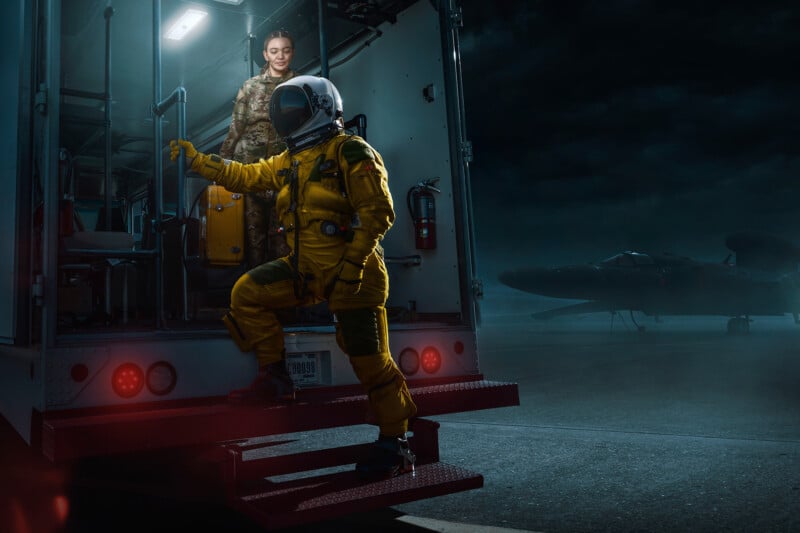 A person in a yellow flight suit steps out of a vehicle while another in military attire observes from behind. It's nighttime, with a dark sky and an aircraft in the background on the tarmac.