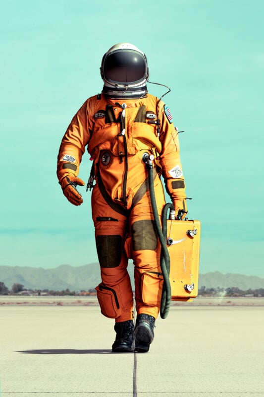 An astronaut in an orange spacesuit walks confidently on a runway, holding a metal case. The background features a clear blue sky and distant mountains.