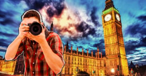 A person in a red plaid shirt is holding a camera and taking a photo in front of the illuminated Big Ben and the Houses of Parliament in London, with a dramatic, cloudy sky at dusk.