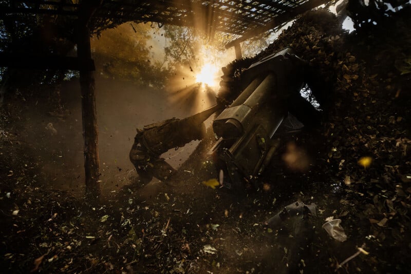 A mechanical excavator arm amidst flying debris and foliage, silhouetted against a sunlit backdrop. Sun rays filter through a lattice roof structure, creating a dramatic contrast between light and shadow.