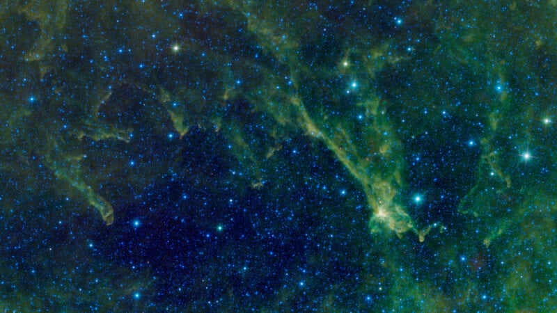 A vibrant image of outer space shows a dense field of blue and green stars shimmering against a deep, dark background. Wispy clouds of luminous green gas and dust stretch across the cosmic scene, creating a mystical, ethereal effect.