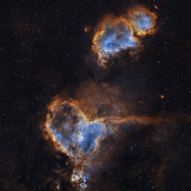 A stunning view of the Heart and Soul Nebulae in deep space, showing intricate formations in blue and orange hues against a backdrop of countless stars. The nebulae resemble cloudy, interconnected regions.