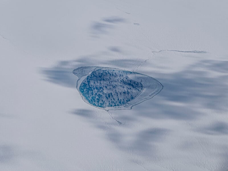 Aerial view of a small, thawed blue lake surrounded by cracked ice and snow. Shadowy patterns stretch across the snowy surface, hinting at the cold, remote setting.