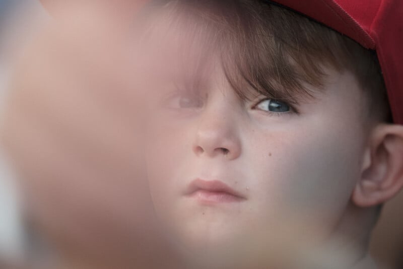 A child wearing a red cap looks through a blurred foreground, with a soft focus on their face, giving an introspective and thoughtful expression.