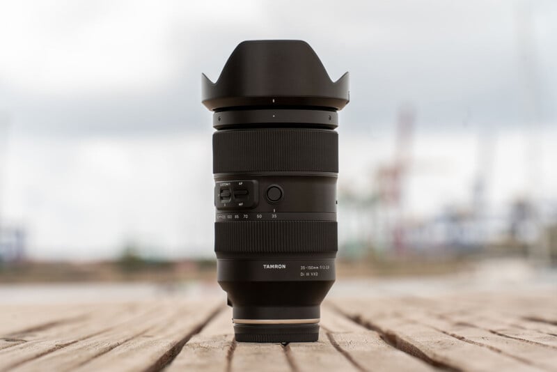 A black Tamron camera lens with a wide-angle hood, standing on a wooden surface against a blurred outdoor background.