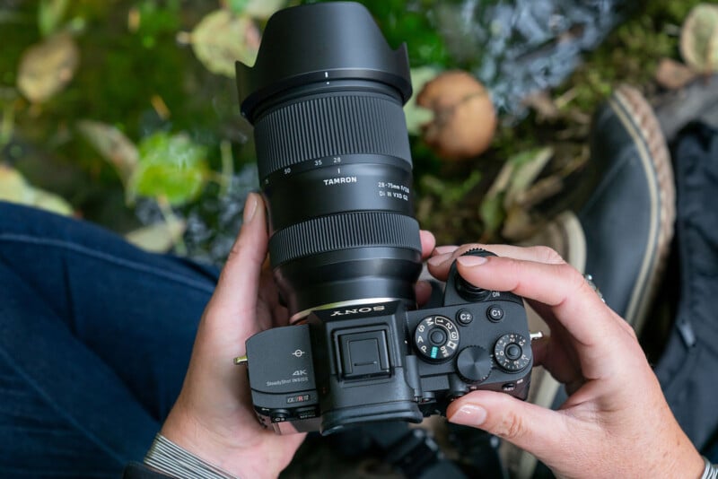 A person holds a Sony camera with a Tamron lens, pointing downward. The backdrop features fallen leaves and blurred water. The person's jeans and shoes are partially visible.