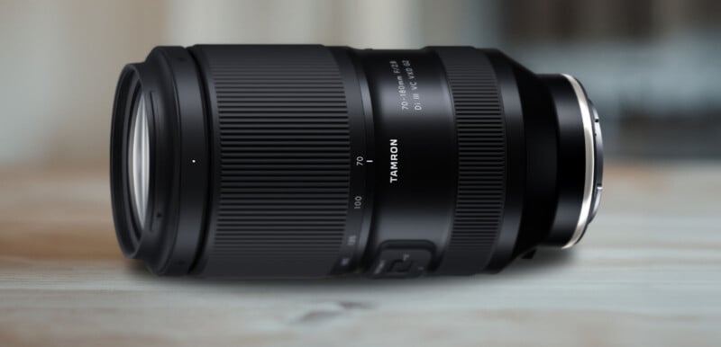 A close-up image of a Tamron 70-180mm f/2.8 Di III VXD G2 camera lens lying on its side on a wooden surface. The lens is black with a ribbed focus ring and silver-colored elements at the mount end.