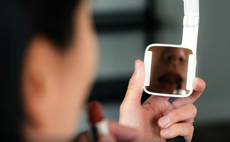 A person applying lipstick is looking into a small, rectangular mirror held in their left hand. The focus is on the reflection of their mouth. The background is blurred.