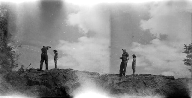 A vintage black and white photo shows four people standing on rocky terrain under a cloudy sky. Two are facing forward on the right, while two are on the left, one pointing to the distance. The image has a faded texture.