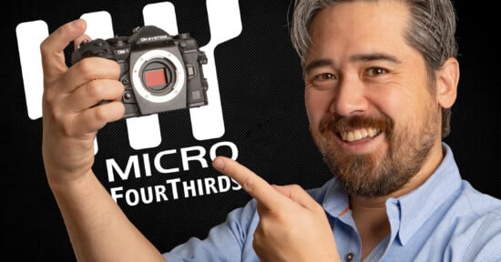 A man smiles and points at a camera he is holding. The camera has no lens attached. Behind him, a logo reads "Micro Four Thirds" on a dark background.