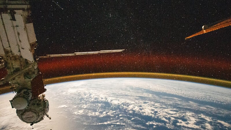 View from space of Earth's curved horizon, showing a bright atmospheric glow in orange and red layers against a star-speckled black sky. Part of a space station is visible in the foreground with Earth’s surface partially covered by clouds.