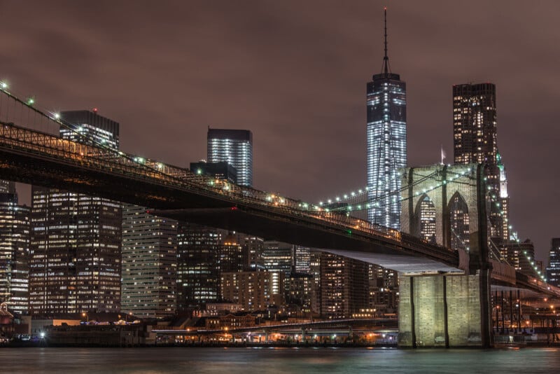 A ⁣nighttime cityscape of New York City featuring the illuminated Brooklyn Bridge in the foreground. Skyscrapers with shining lights stand prominently, including the One World Trade ‍Center ⁢tower, against a cloudy night sky.