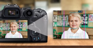 A digital camera on a table, showcasing facial detection technology. The camera screen displays a young girl in focus within a classroom, with lines highlighting facial features. The background shows blurred educational posters.