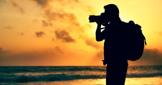 Silhouette of a person with a backpack taking a photo at the beach during sunset. The sky is filled with orange and yellow hues, and the ocean waves gently roll onto the shore.