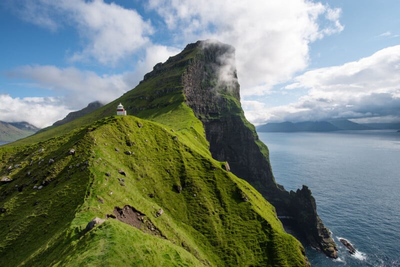 A scenic view of a lighthouse perched on ‌a grassy cliff‌ overlooking the ocean. The rugged green terrain leads to a majestic, cloud-topped mountain.