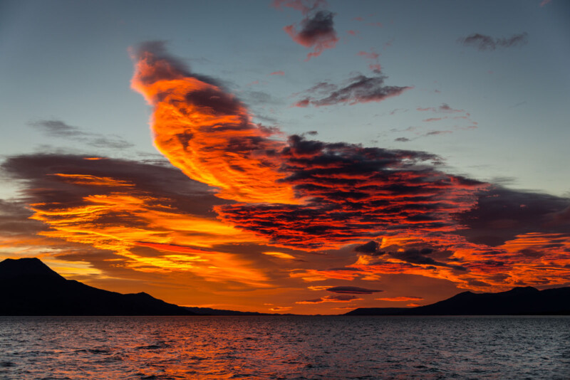 A stunning sunset over a calm body of water, with ⁤dramatic orange and red clouds filling the sky. silhouettes of distant mountains frame ‌the ⁤horizon, creating a breathtaking landscape.