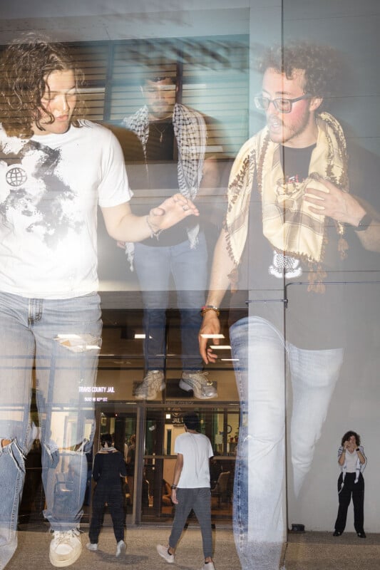 Two men walk outside a building at night, wearing casual clothes and scarves. The scene is a double exposure, blending them with a background of a lit doorway and people entering. One man holds a phone and the other holds a cigarette.