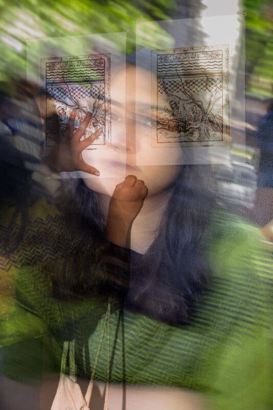 A woman in a green sweater appears in a double exposure image, standing in front of two framed drawings. Her hand is raised, touching one of the frames. The background is blurred with green and brown tones, creating an ethereal effect.