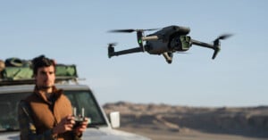 A person standing next to a vehicle, flying a drone in a desert landscape. The person holds a remote control, and the sky and distant hills are visible in the background.