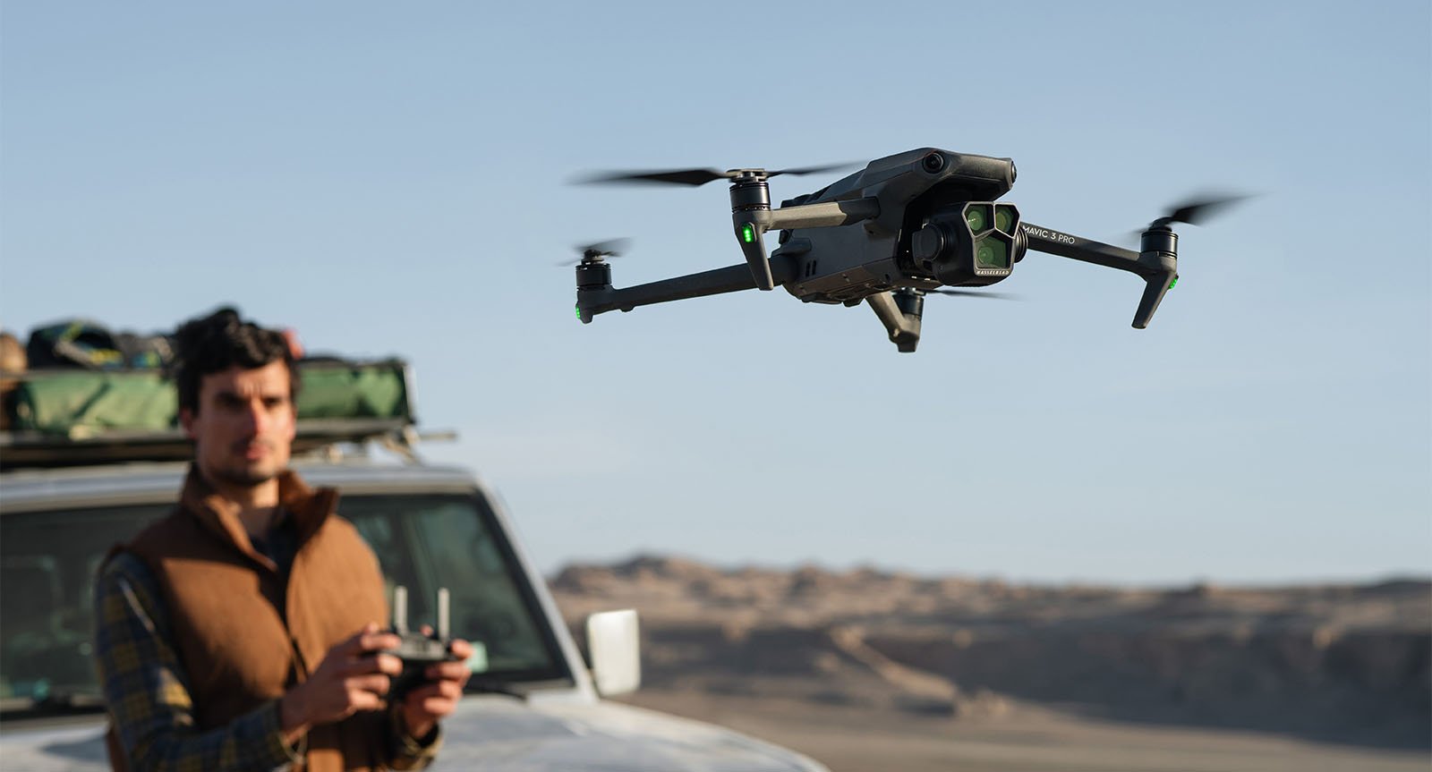 A person standing next to a vehicle, flying a drone in a desert landscape. The person holds a remote control, and the sky and distant hills are visible in the background.