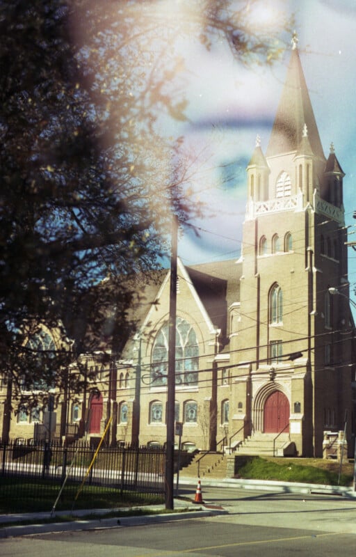 A large brick church with a tall steeple and red doors stands behind a street. The building features arched windows and ornate architectural details. Surrounding trees and power lines are visible under a clear blue sky.