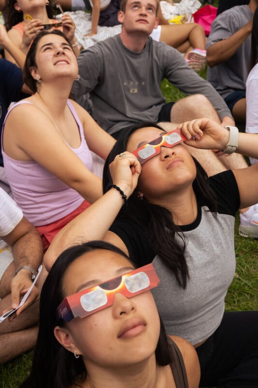 A group of people sitting on grass, wearing special glasses and looking up at the sky, likely observing a solar eclipse. The scene conveys a sense of curiosity and wonder.