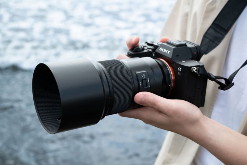 A person holding a Sony Alpha 7R camera with a large telephoto lens, standing near a body of water. The camera strap is visible around their neck, and the background features blurred waves or water texture.