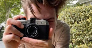 A person holding a vintage camera up to their face, taking a photo. The background features greenery with bushes and trees. The person's hair is windswept and they are wearing a beige shirt.