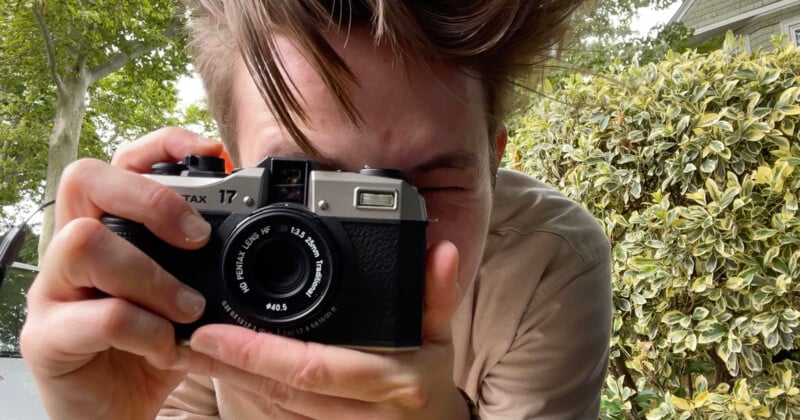 A person holding a vintage camera up to their face, taking a photo. The background features greenery with bushes and trees. The person's hair is windswept and they are wearing a beige shirt.