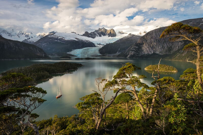 Scenic view of a​ valley with a calm lake surrounded by ​lush green trees and rocky ‍hills. In the background, a glacier rests between snow-capped mountains under​ a partly cloudy ⁤sky. A sailboat is anchored in the lake.