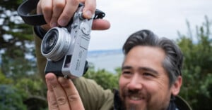 A person smiles while holding up a Fujifilm camera outdoors, with greenery and a blurred landscape in the background.