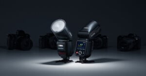 Studio lighting equipment, including two Godox flash units with digital displays, posed against a dark background. In the distance, silhouettes of cameras are visible.