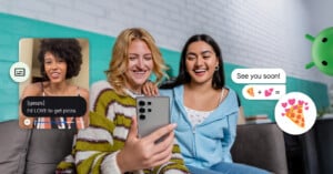 Three friends sit on a couch, smiling as they look at a smartphone. A video call shows a woman saying she loves pizza. Chat bubbles display a pizza emoji and hearts. The setting is casual with a light, brick wall background.