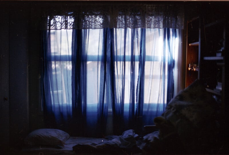 A dimly lit room with a window covered by sheer blue curtains and lace. Soft light filters through, casting shadows on a messy bed with pillows in the foreground. A bookshelf is partially visible to the right.